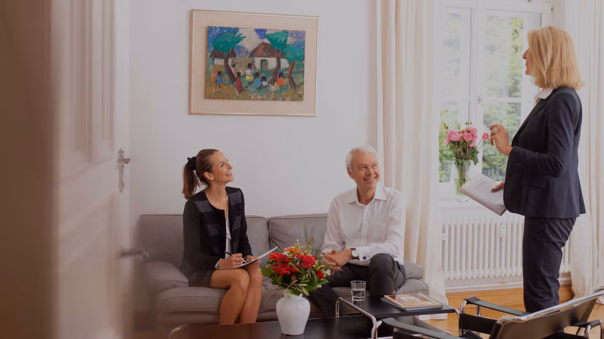 Mittig sitzen zwei Personen auf einem grauen Dreisitzer. Die linke Person ist eine Frau mit dunklen mittellangen Haaren, die zu einem Pferdeschwanz zusammengebunden sind. Die ist die Ansprechpartnerin für das Recruiting - Magdalena Kalus. Sie trägt eine schwarze Jacke und darunter eine weiße Bluse. Neben ihr sitzt ein älterer Herr - Geschäftsführer der FORUM Burkhard Wittek - mit weißen Hemd. Beide lächeln die Dame - Eva Wunderer aus dem HR - an. Sie steht auf der rechten Seite des Bildes. Die Dame auf der rechten Seite des Bildes trägt einen dunkelblauen Hosenanzug mit offenem Blazer und hält einen Notizblock in der Hand. Im Hintergrund oberhalb der Couch hängt ein Bild an der Wand. Auf dem Fensterbrett rechts neben der Couch steht eine Vase mit rosafarbenen Chrysanthemen. Am linken Rand des Bildes ist die Tür noch angeschnitten. Hierdurch erkennt man, dass das Bild in den Raum hinein aufgenommen wurde.